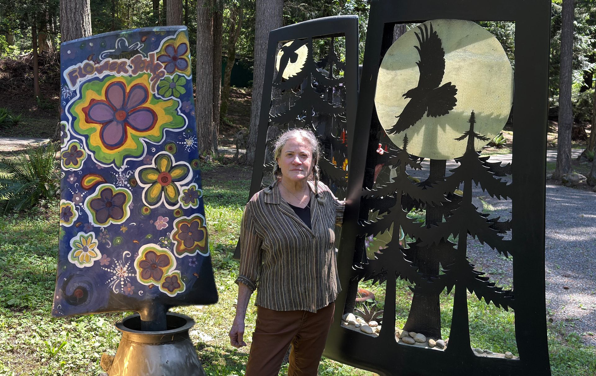 Jill Drllevich standing between her Flower Folk painted panel and a Breathe Deep raven sculpture in a forest clearing